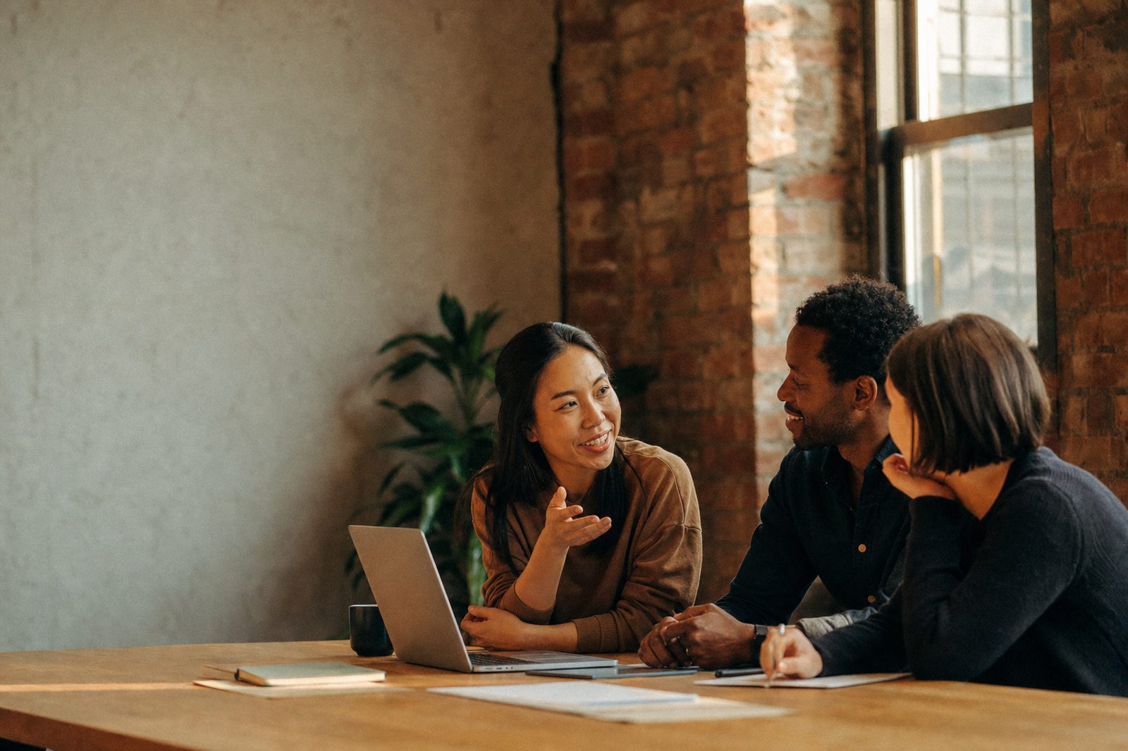 Three colleagues in conversation around a table with a laptop and notebooks in a warmly lit office